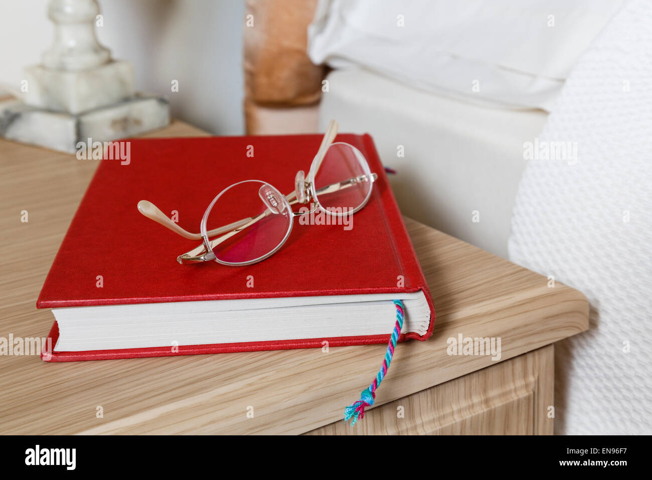Generische einfache rote Hardback Buch und Lesebrille auf einem Nachttisch an der Seite eines Bettes in einem häuslichen Schlafzimmer zu Hause. England, Großbritannien Stockfoto