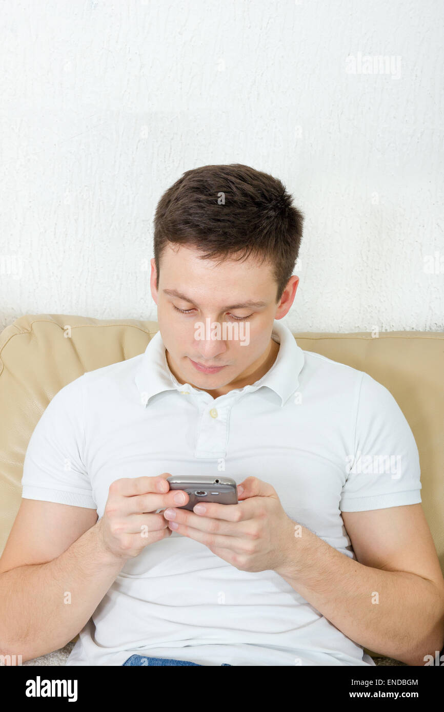 Junger Mann mit seinem mobilen Smartphone senden, glücklicher Kerl sitzt auf der Couch SMS auf Handy zu Hause Stockfoto