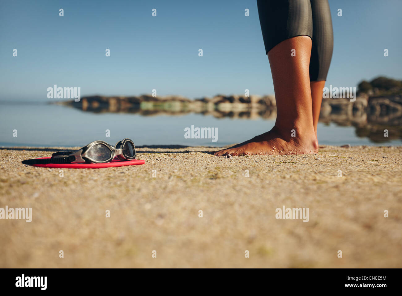 Schwimmen-Schutzbrillen auf dem Sand mit Füßen einer Frau stehen. Brille im Vordergrund. Stockfoto