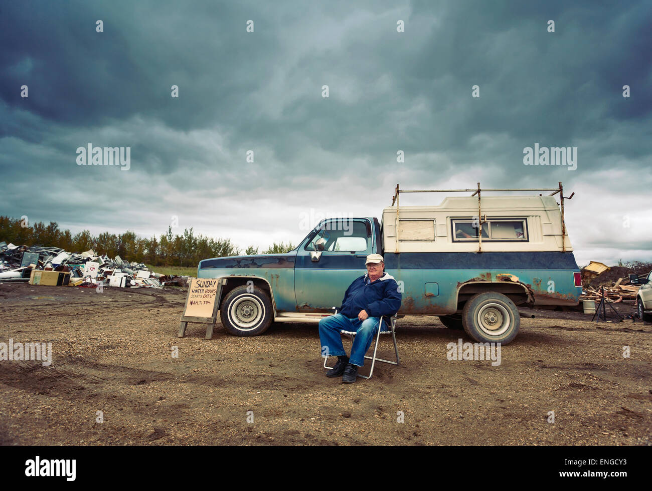 Ein reifer Mann sitzt auf einem Stuhl seinen Pick up Truck. Berge von Müll, Schrott, Metall und Holz-Objekte. Stockfoto