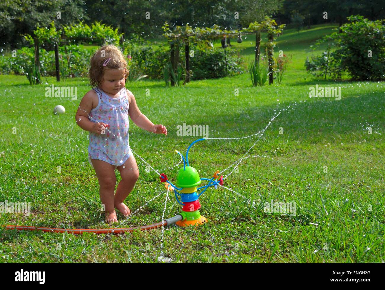 Blonden lockigen Haaren nass Kleinkind spielen mit einem Spielzeug-Sprinkler.  Wassertröpfchen sprühen rund um. Stockfoto