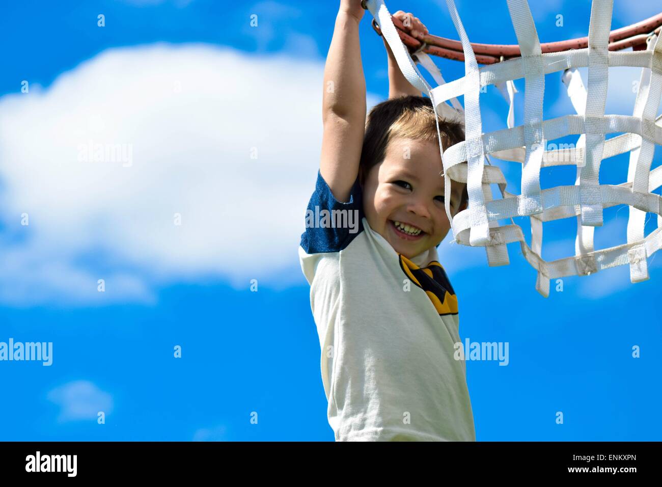 3 Jahre alter Junge mit großen braunen Augen haltend auf einen Basketballkorb mit den blauen Himmel hinter ihm lächelnd. Stockfoto
