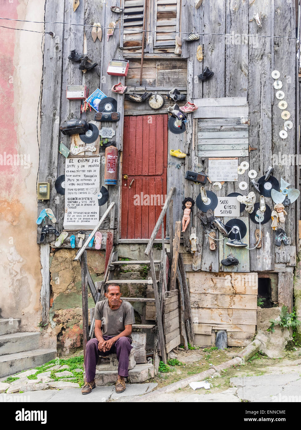 Ein Hispanic kubanischen sitzt Man auf den Stufen von seinem sehr seltsam, eingerichtete unkonventionell Holzhaus. Stockfoto