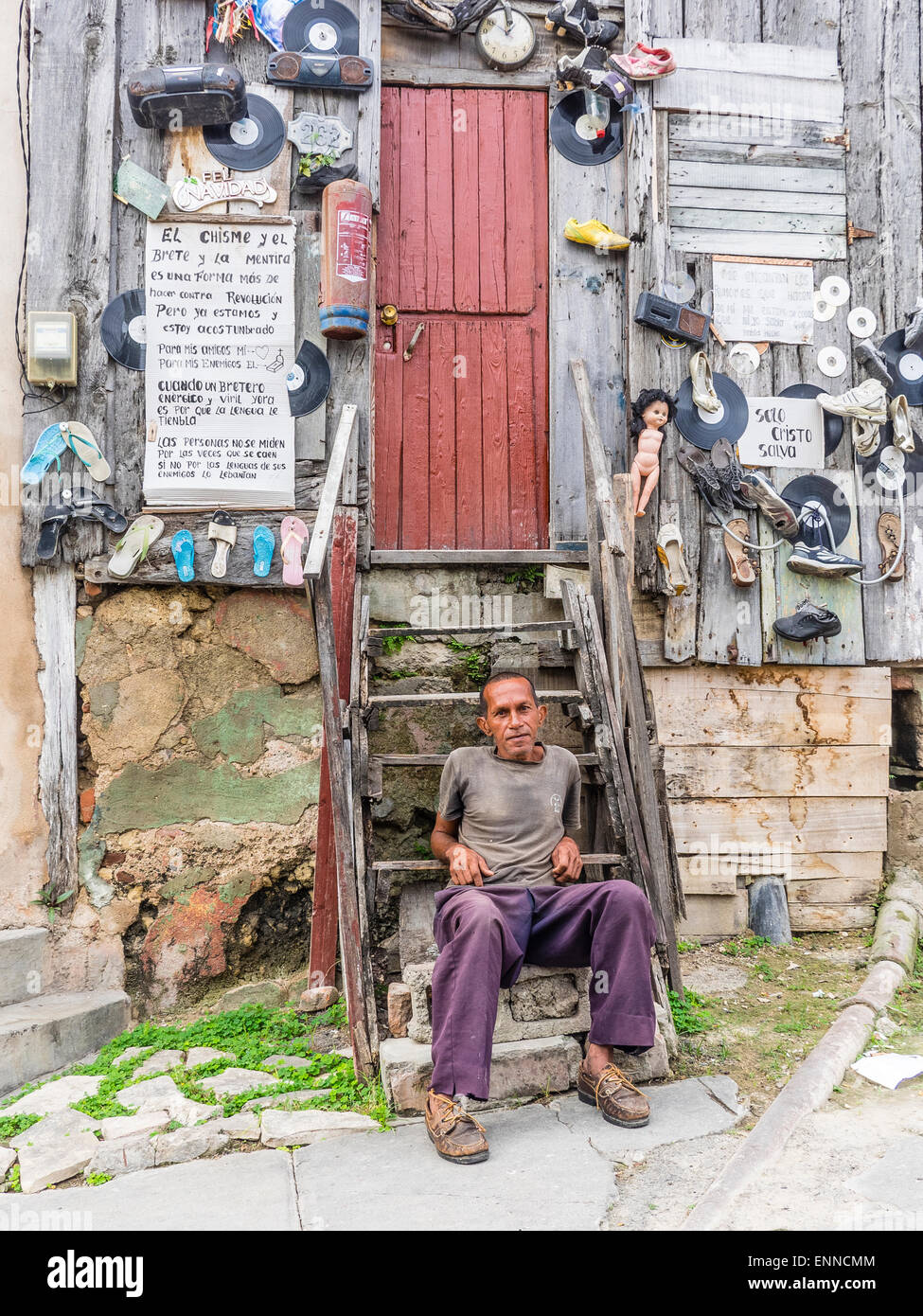 Ein Hispanic kubanischen sitzt Man auf den Stufen von seinem sehr seltsam, eingerichtete unkonventionell Holzhaus. Stockfoto