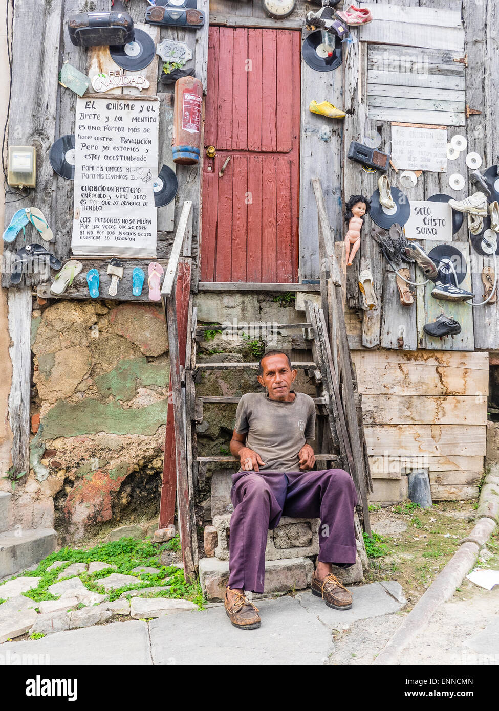 Ein Hispanic kubanischen sitzt Man auf den Stufen von seinem sehr seltsam, eingerichtete unkonventionell Holzhaus. Stockfoto