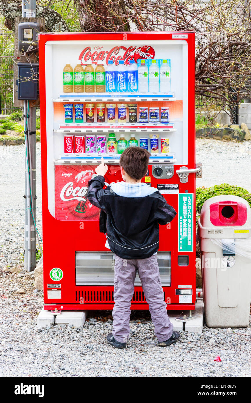 Rückseite des kaukasischen Kindes, Junge, 8-9 Jahre alt, stehend vor dem japanischen Coca-Cola-Automaten, der über ihm aufragt und Knopf für ein Getränk drückt. Stockfoto