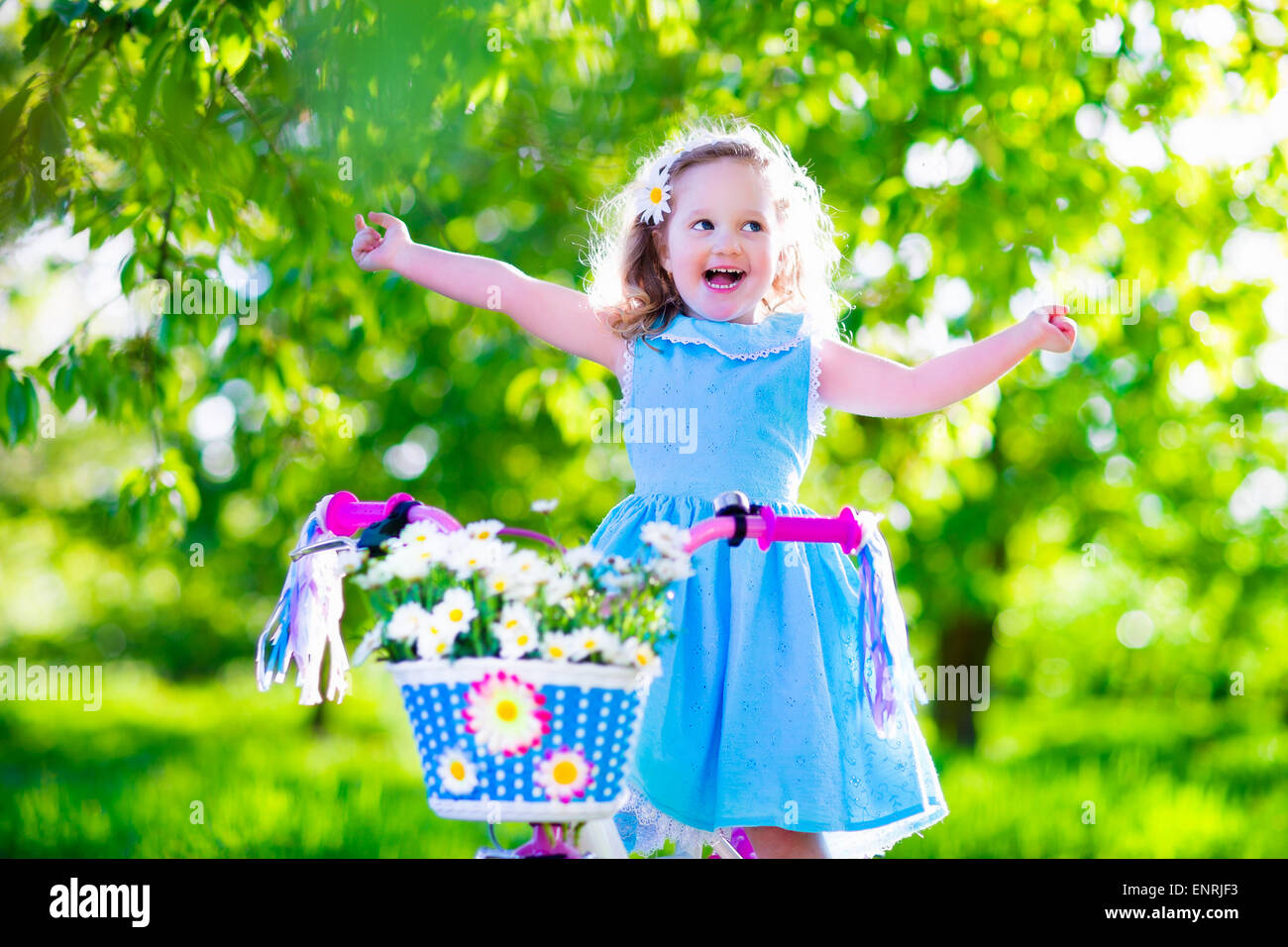 Glückliches Kind mit dem Fahrrad. Süßes Kind Radfahren im Freien. Kleines Mädchen in einem blauen Kleid auf einem rosa Fahrrad mit Blumen-Gänseblümchen Stockfoto