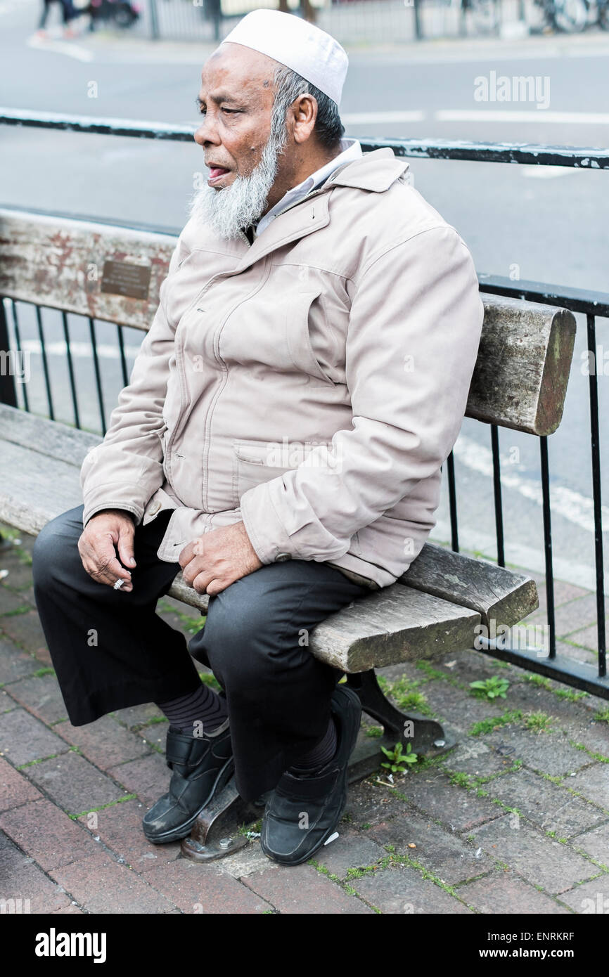 Asiatische tragen Topi Kopf saß in Salisbury Street Wear Rauchen einer Zigarette auf der Straße Sitzbank Stockfoto