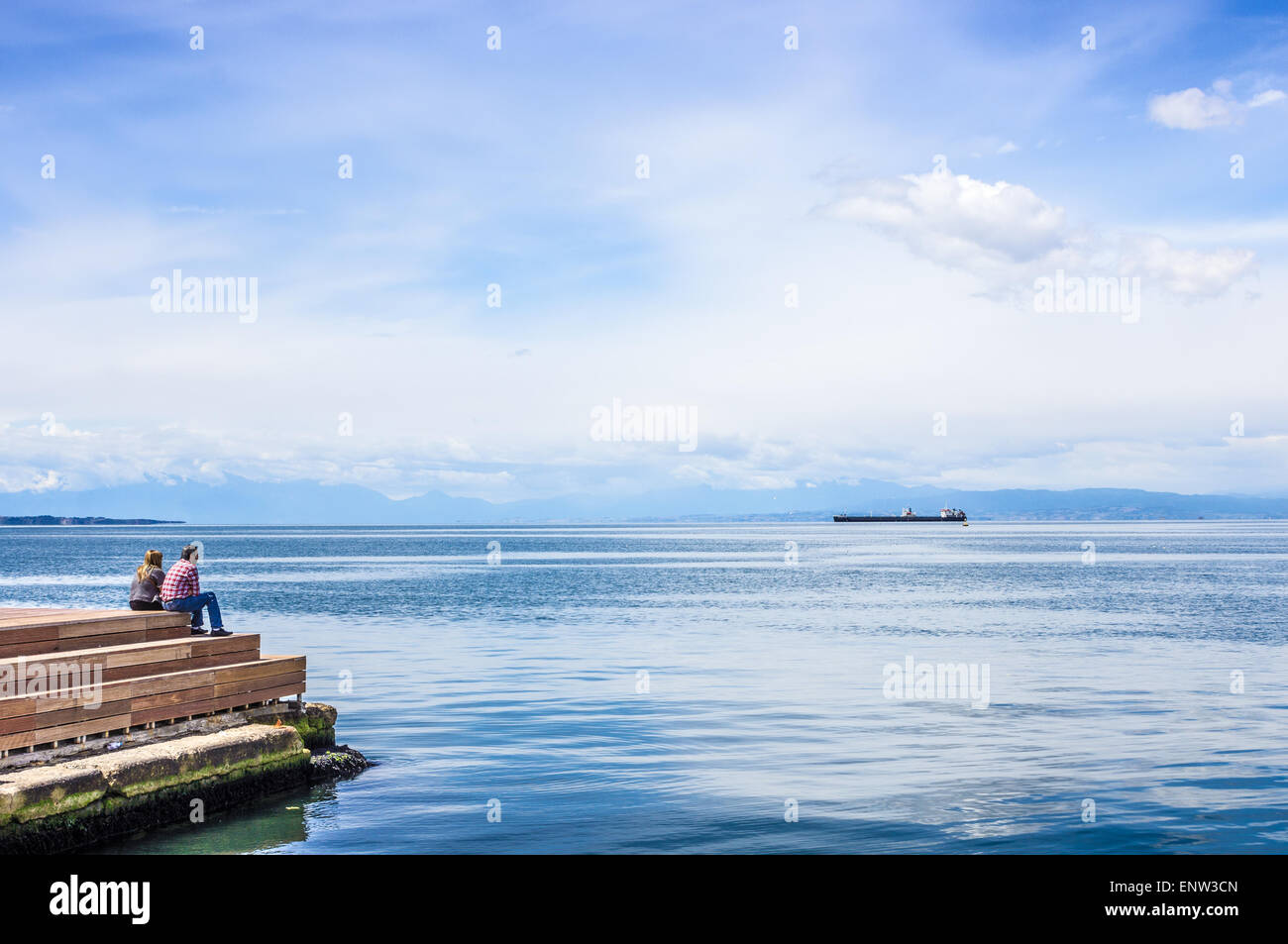 Paar mittleren Alters sitzen auf Pier vor Tanker auf das tiefblaue Meer und mythologischen Mount Olympus am Horizont Stockfoto