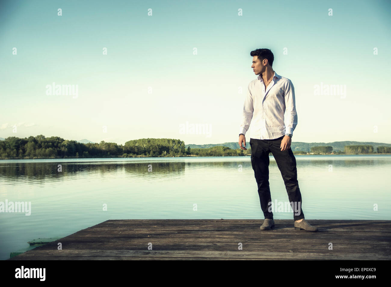 Hübscher junger Mann an einem See an einem sonnigen, ruhigen Tag, stehend auf einem hölzernen pier Stockfoto