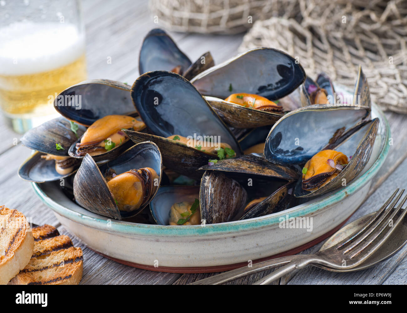 Eine Schüssel mit leckeren gedämpfte Muscheln mit gegrilltem Brot und Bier auf einem rustikalen Tisch mit Fischnetz. Stockfoto