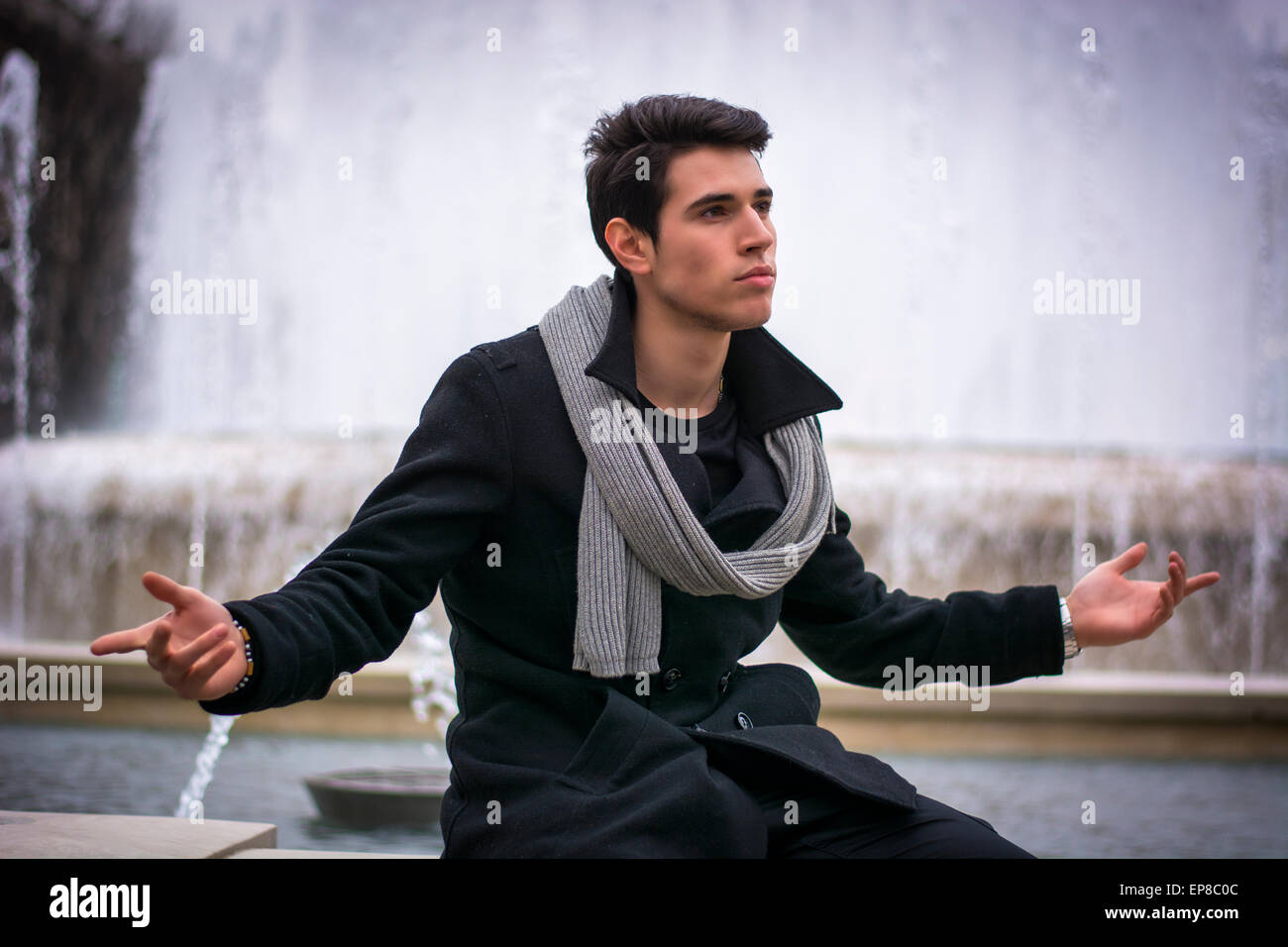 Junger Mann draußen sitzt neben großer Brunnen mit Armen verteilt offen und selbstbewusst Ausdruck Stockfoto