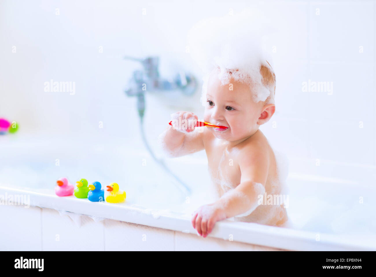 Niedlichen kleinen Jungen seine Zähne putzen, spielen mit Schaum und bunte Kautschuk Ente Spielzeug in einem weißen sonnigen Bad Bad Stockfoto