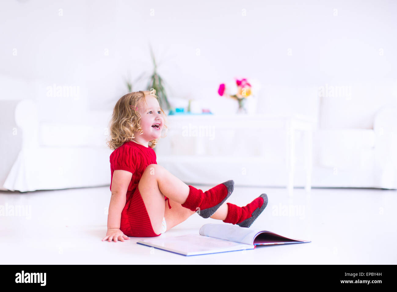 Niedliche kleine Mädchen mit dem lockigen Haar mit einer warmen roten Strickkleid und Socken ein Buch auf dem Boden sitzend Stockfoto