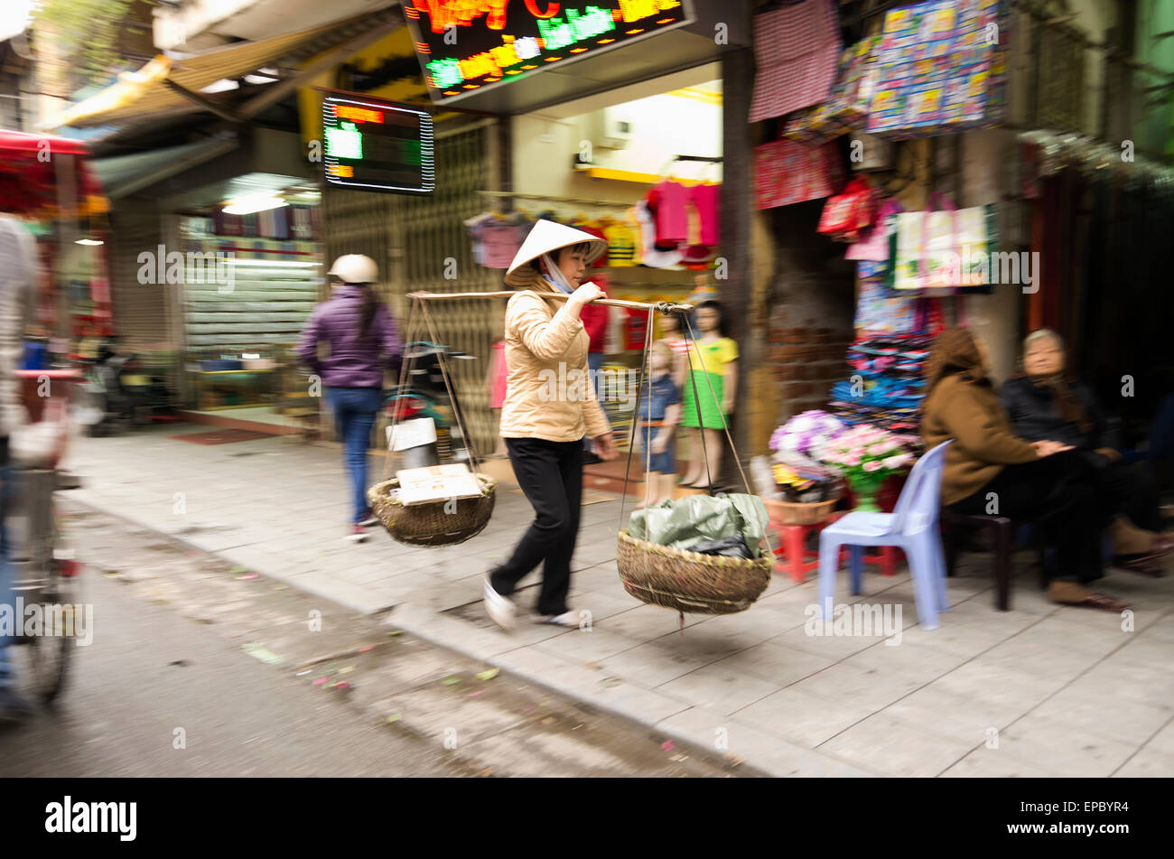 Weibliche Straßenhändler trägt zwei Körbe von verschiedenen Dingen in Körben an jedem Ende des Pols ausgesetzt; Hanoi, Vietnam Stockfoto