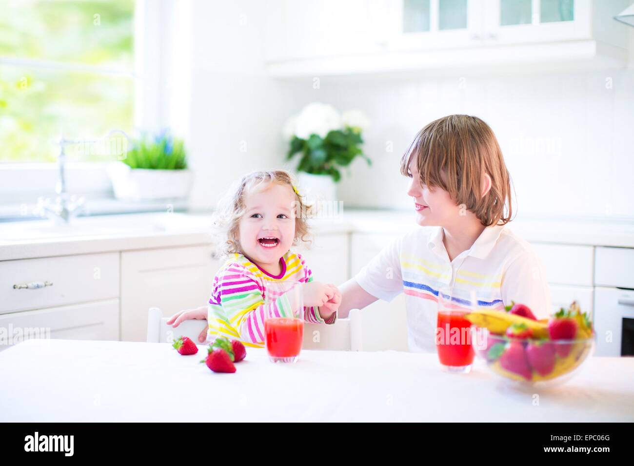 Glücklich Teenager-jungen und seine niedlichen Kleinkind Schwester mit Obst zum Frühstück trinken Saft in einer sonnigen weißen Küche Stockfoto