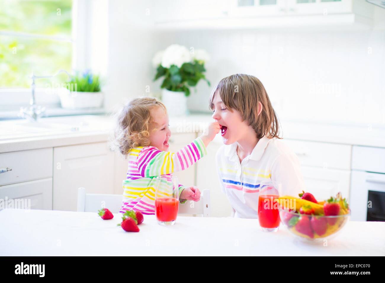 Glücklich Teenager-jungen und seine niedlichen Kleinkind Schwester mit Obst zum Frühstück vor der Schule in einer sonnigen weißen Küche Stockfoto