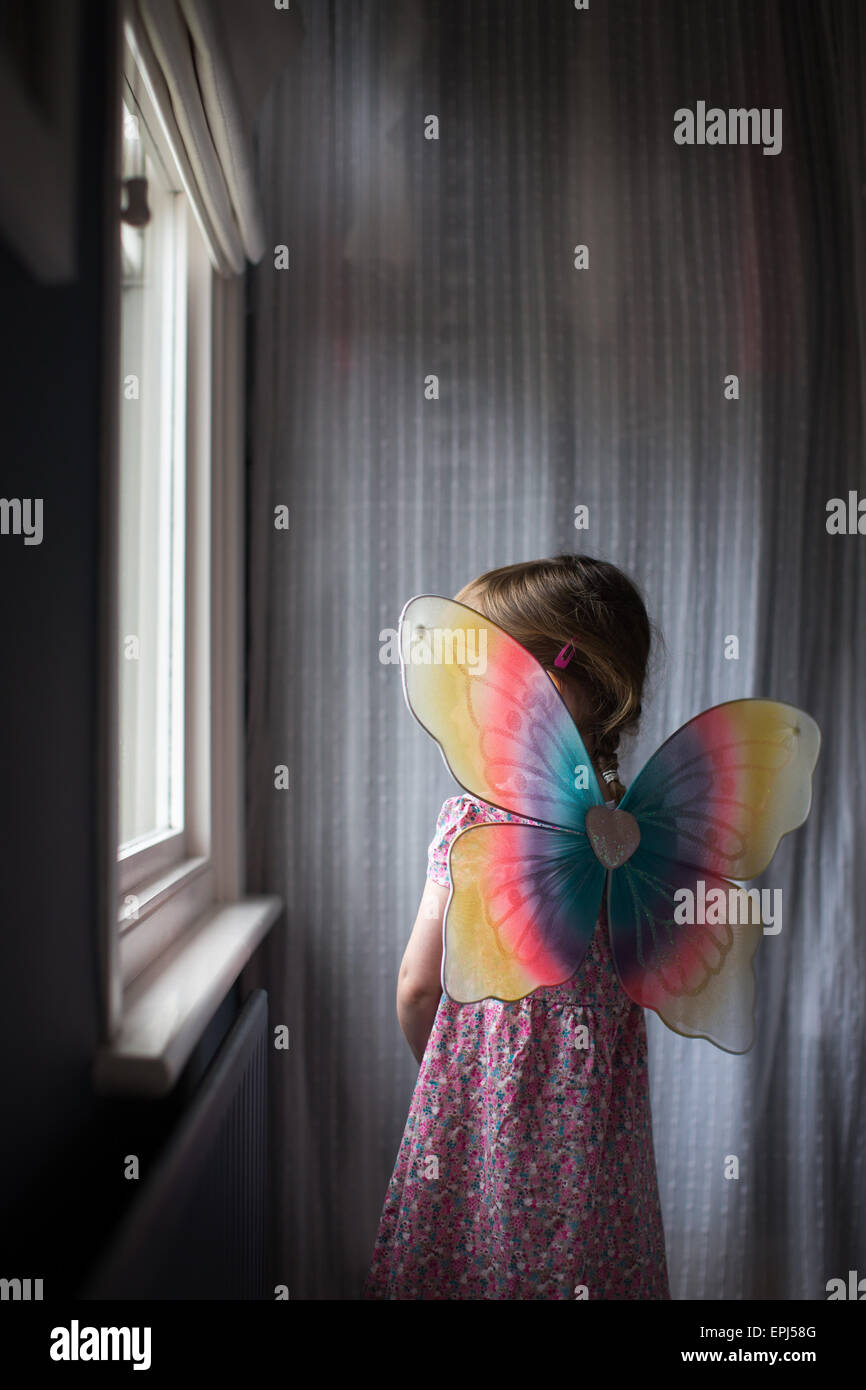 Ein Kind im Alter von 3 sieht aus dem Schlafzimmerfenster tragen bunte Feenflügel Stockfoto