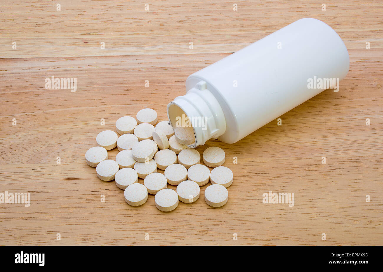 Vitamintabletten aus einer weißen Flasche geschüttet und gezeigt auf einem hölzernen Hintergrund Stockfoto
