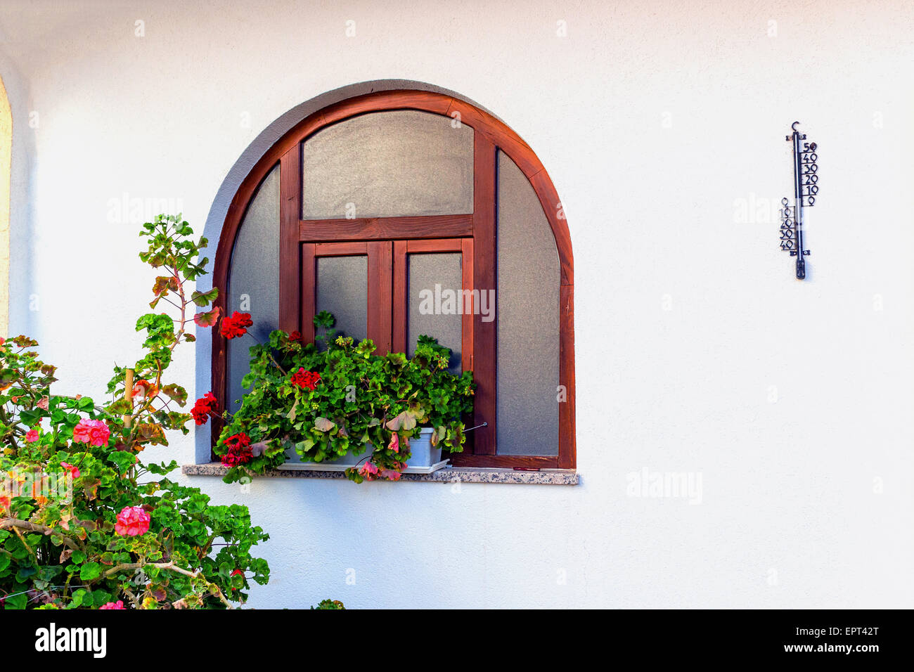 Abgerundete Holzrahmen Fenster mit roten Geranien Blumentöpfe und schwarzem Eisen Thermometer auf weißen Wand Stockfoto