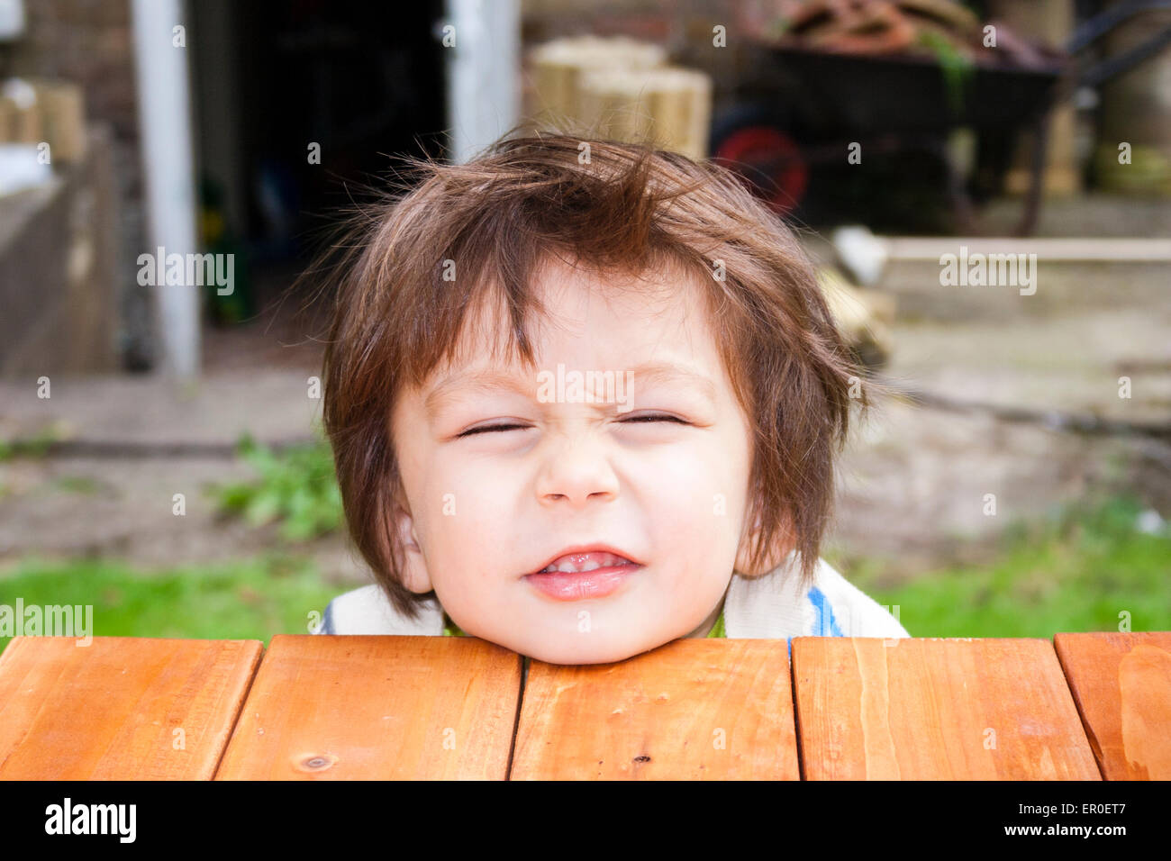 Nahaufnahme des Gesichts eines kleinen Kindes, Junge, 3-4 Jahre alt, während er das Kinn auf einer Holzplattform aufliegt, dem Betrachter zugewandt und sein Gesicht nach oben schraubt. Im Freien Stockfoto