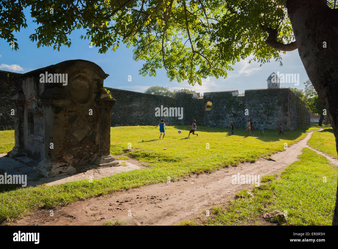 Kinder, die Fußball auf einem grasbedeckten Feld in der Nähe der Außenwand von Fort Speelwijk spielen, wo ein europäischer Grabstein in Old Banten, Banten, Indonesien, liegt. Stockfoto