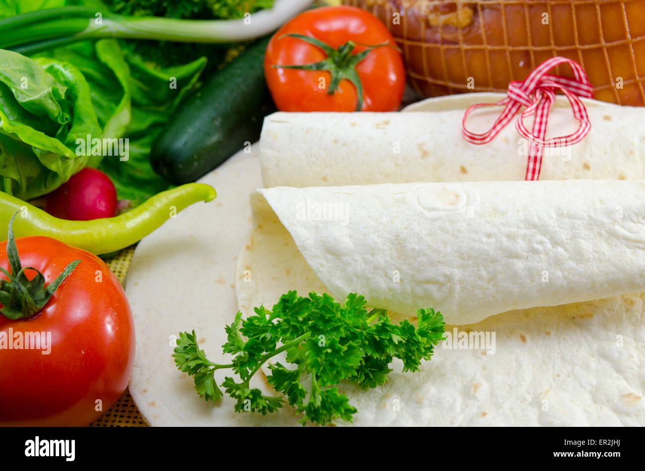 Leere Tortillas gebunden mit einem roten Band auf einem Tisch mit Tomaten, Salat und Schinken Stockfoto