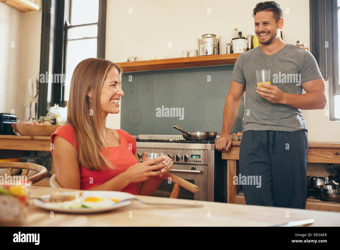 Aufnahme des jungen Paares in Küche lächelnd. Junge Frau sitzt am Frühstückstisch mit Mann, die Küchentheke hält eine g Stockfoto
