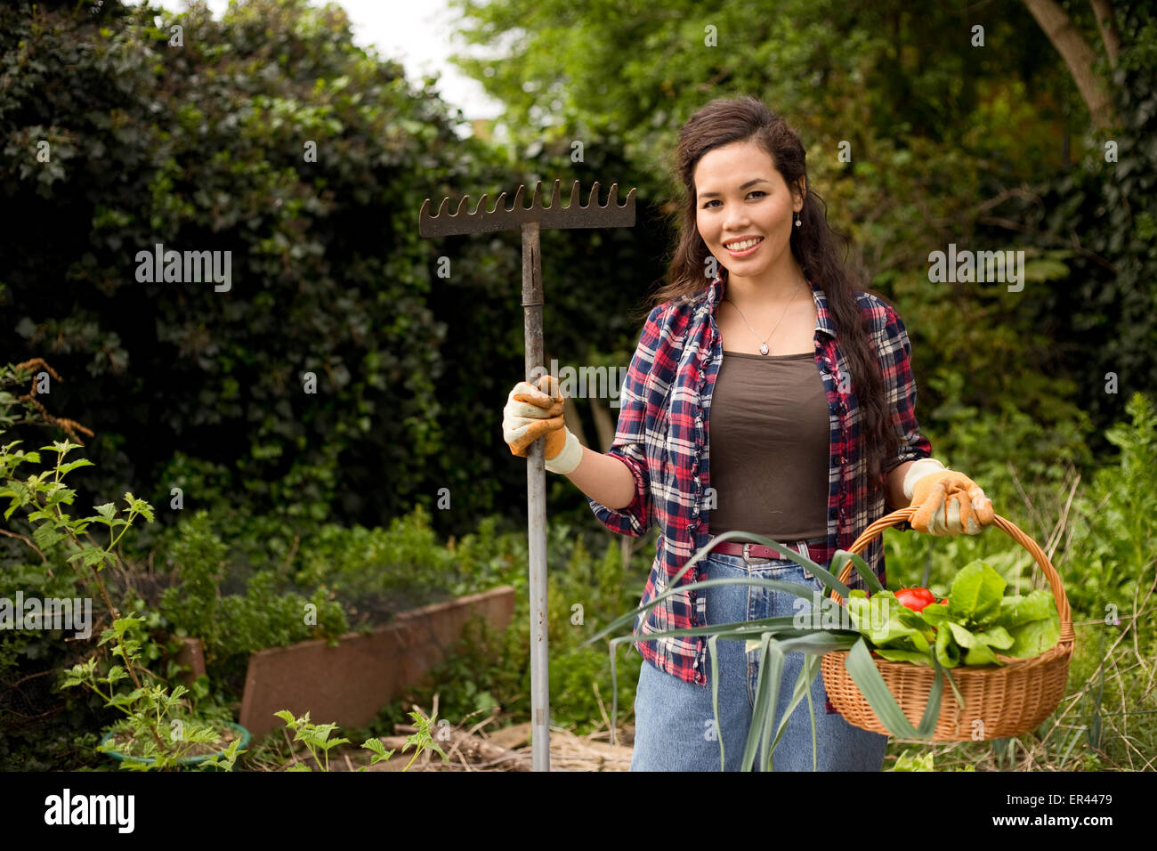 junge Frau im Garten hält einen Korb mit frischem Gemüse und einer Harke Stockfoto