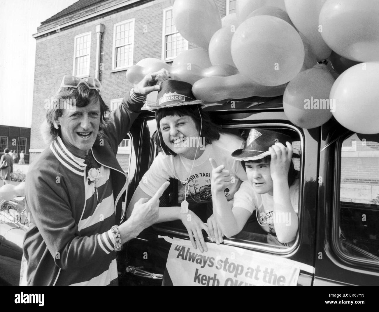 Jimmy Savile versucht den Hut auf der Rosemary Messenger, von Patricia Ecclewaite, beide aus Swinton Krankenhaus beobachtet, während sie darauf, für den "Startschuss" nach Blackpool in der Manchester Taxifahrer Jahresausflug für die Behinderten 21. Juli 1975 warten Stockfoto