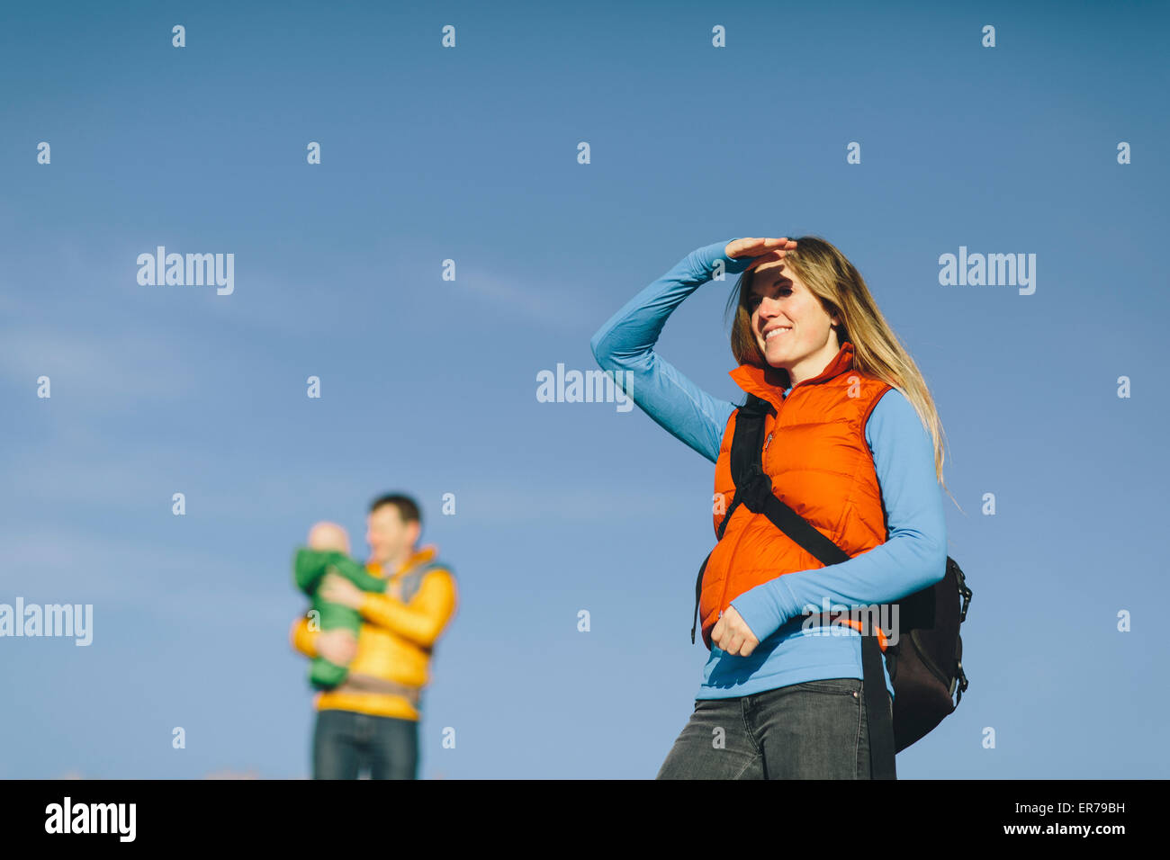 Eine junge Frau beschattet Augen gegen Sonnenuntergang mit Baby und Mann im Hintergrund von einer Winterwanderung. Stockfoto