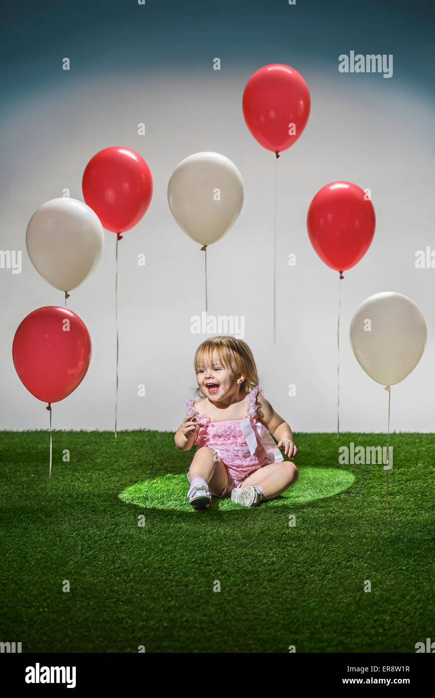 Fröhliches Mädchen sitzen auf dem Rasen mit roten und weißen Luftballons in der Luft Stockfoto