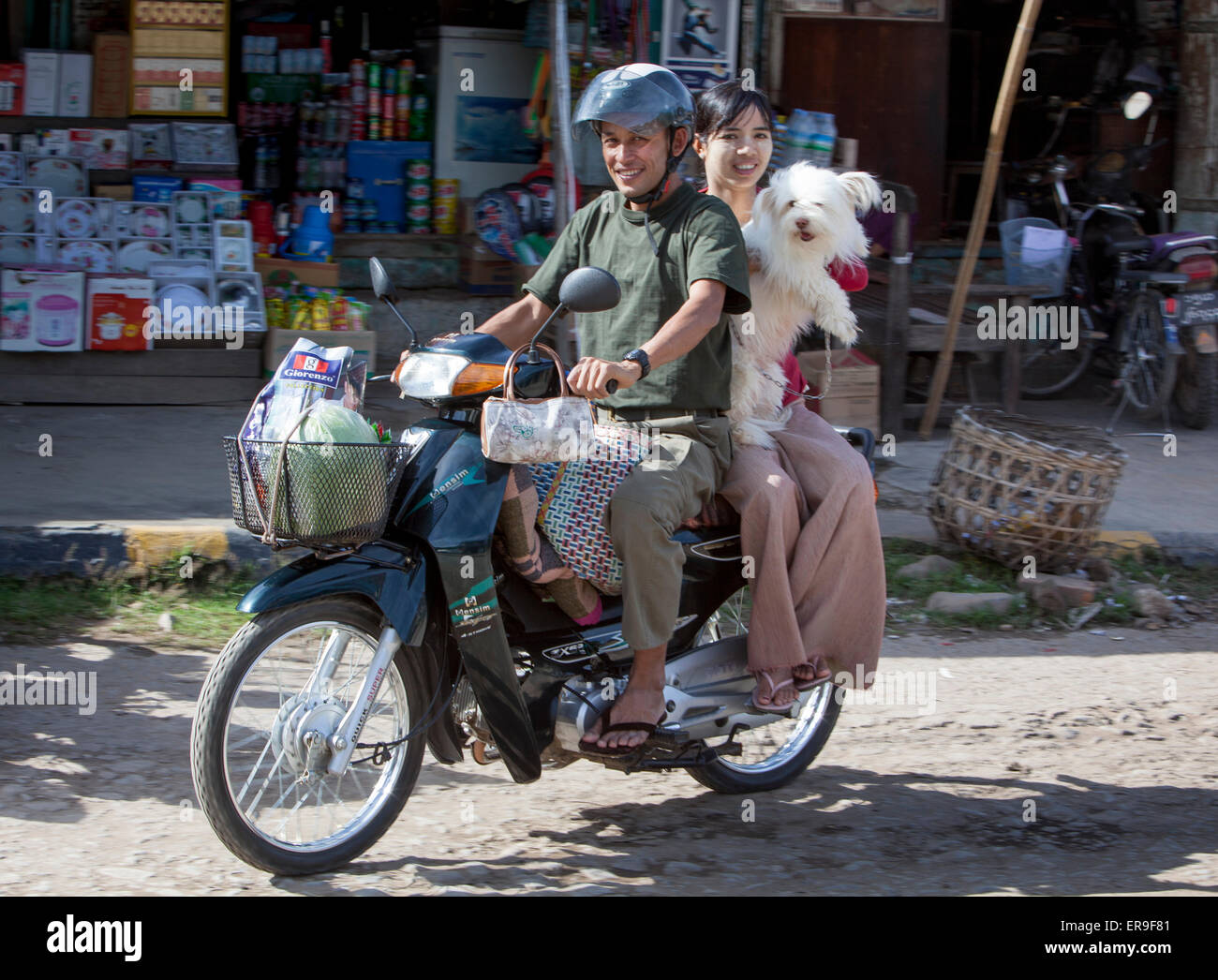 Ein Mann reitet auf seinem Motorrad in der birmanischen Stadt Hsipaw. Seine Frau Passagier hält einen großen weißen Hund. Stockfoto