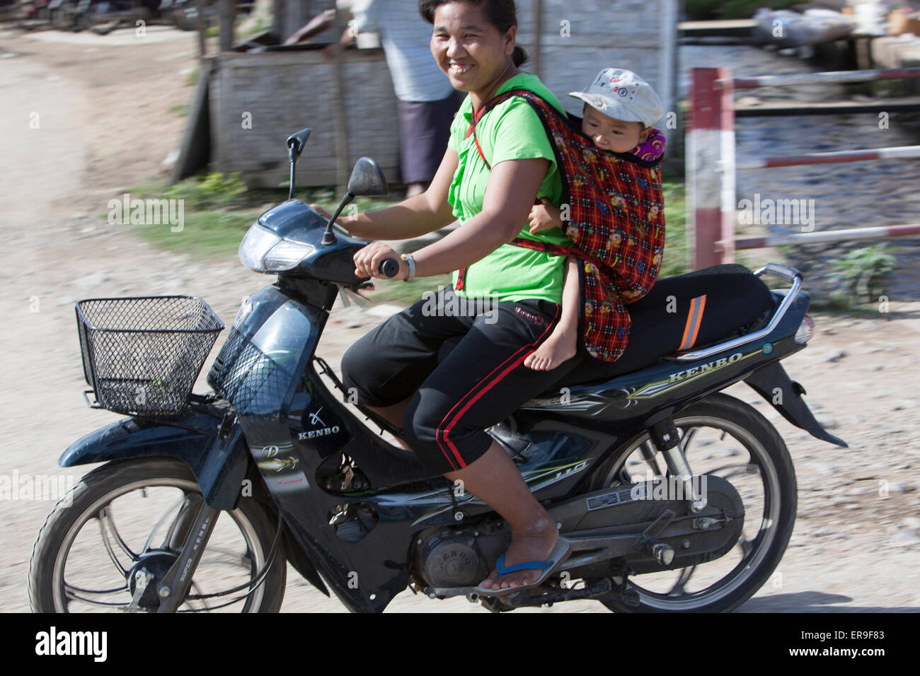 Eine Frau auf einem Motorrad lächelt in die Kamera, als sie vorbei reitet. Ihr Kind ist auf dem Rücken in einem Tuch Riemen geschleudert. Stockfoto