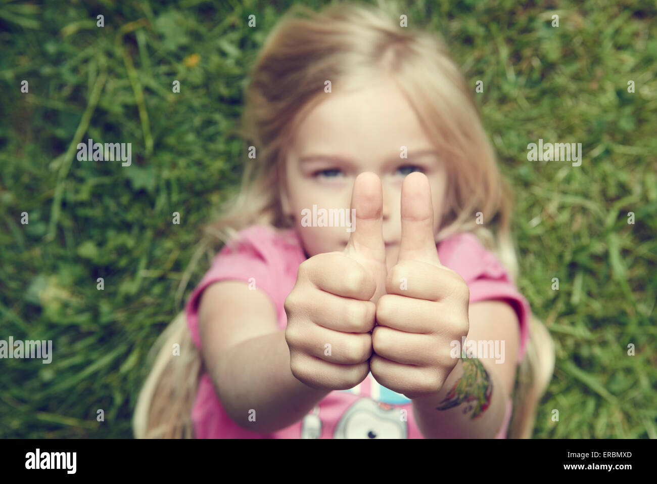 Kind blonde Mädchen auf dem grünen Rasen im Sommerpark mit Daumen hoch Geste mit beiden Hand liegend Stockfoto