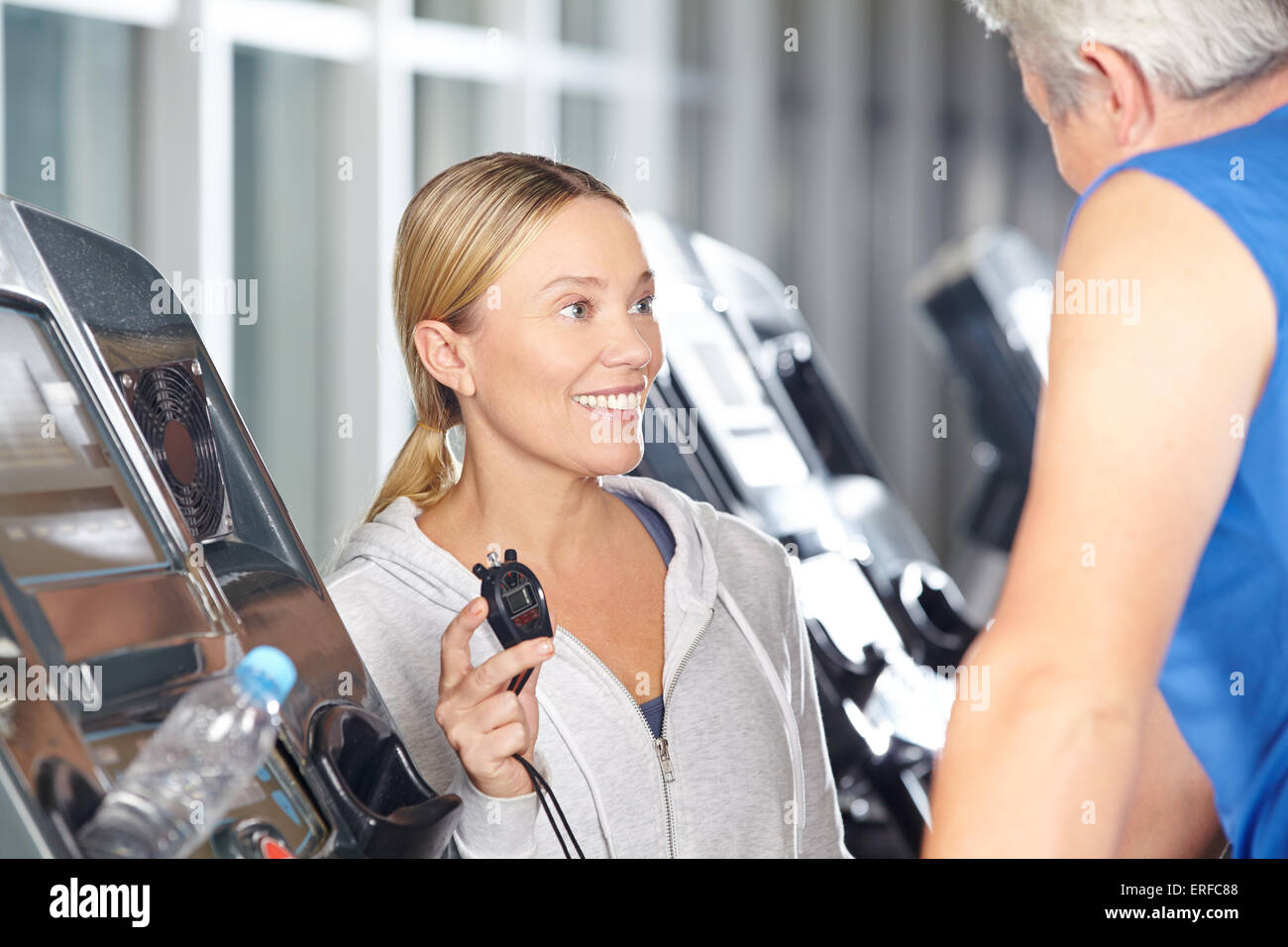 Trainer mit Stoppuhr in einem Fitness-Center im Gespräch mit Senior auf Laufband Stockfoto