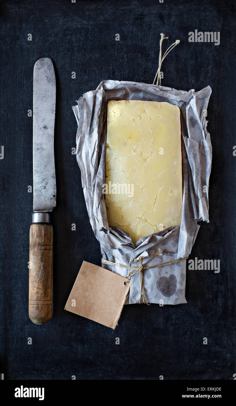 Ein Block von englischen Reifen Cheddar-Käse in ein leeres Etikett mit einem Vintage Messer auf einer Tafel rustikalen Papier eingewickelt. Stockfoto