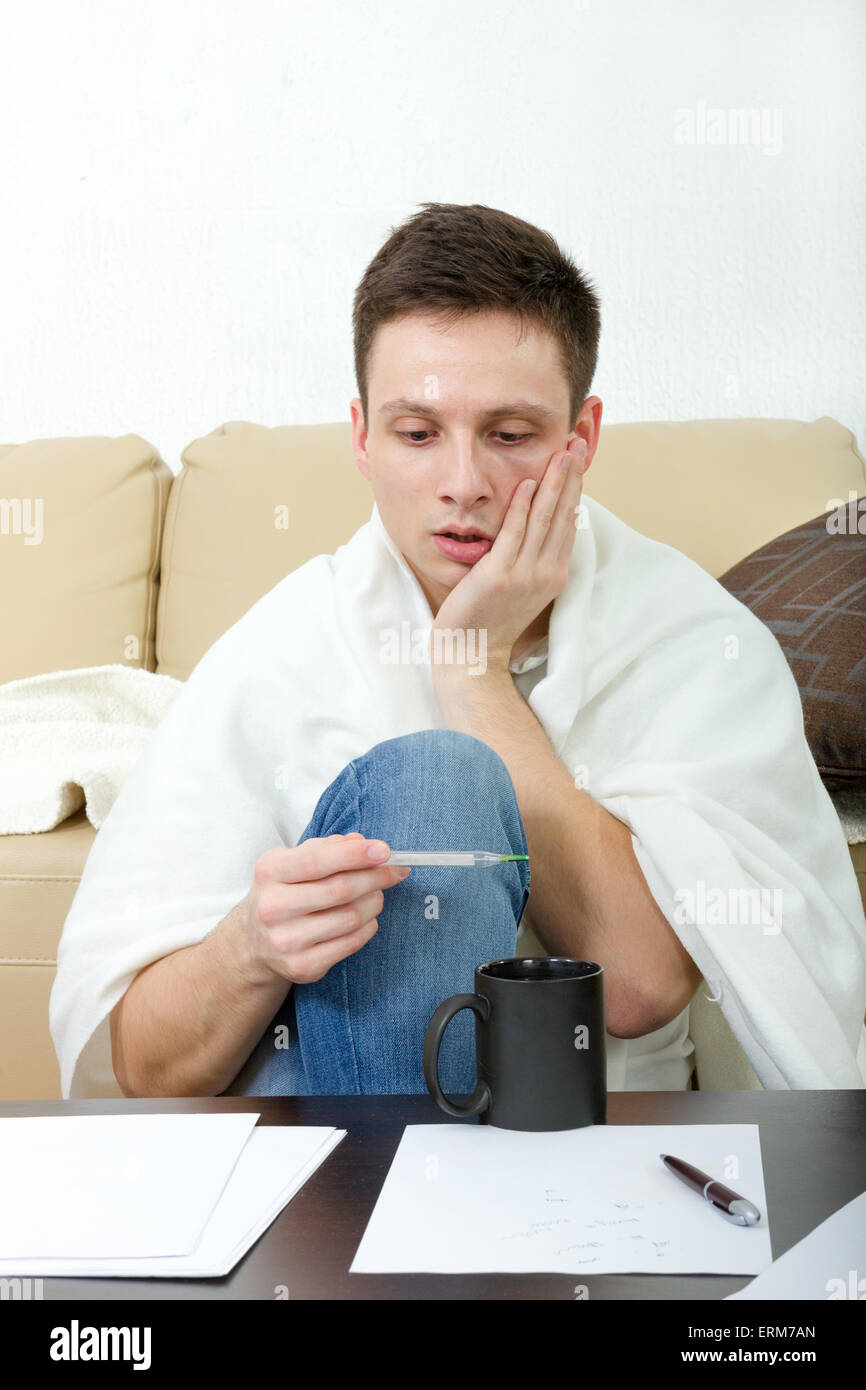 Kranker Mann Temperaturmessung am Thermometer beim Sitzen auf dem Sofa zu Hause. Krank männlichen Studenten mit Kopfschmerzen mit Whit bedeckt Stockfoto