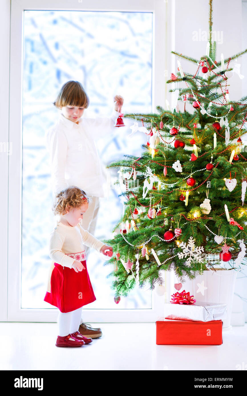 Glückliche Kinder spielen neben einem Weihnachtsbaum. Schöne lockige Kleinkind Mädchen helfen, ihren Bruder, den Weihnachtsbaum zu schmücken Stockfoto