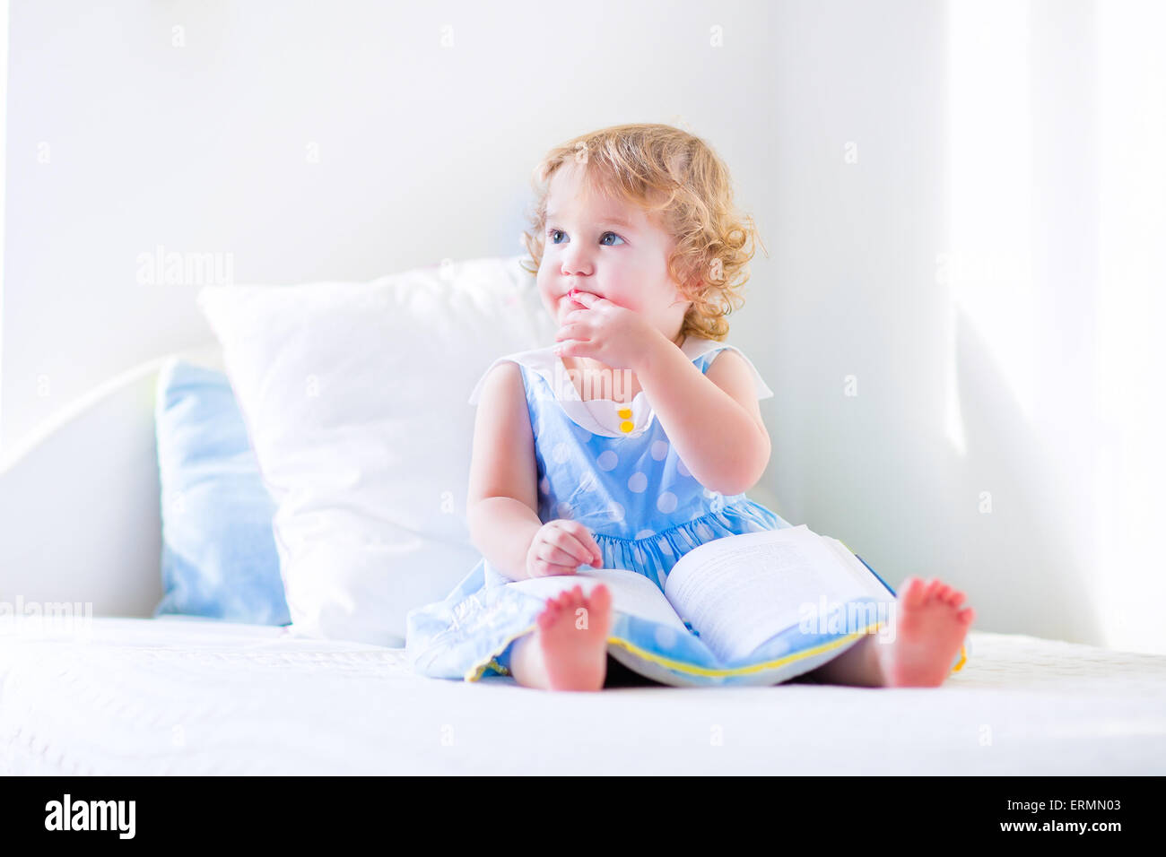 Niedlichen Kleinkind, entzückende lockige kleines Mädchen in einem blauen Kleid, sitzt auf einem Bett, einem Buch spielen in einem sonnigen weiße Schlafzimmer Stockfoto