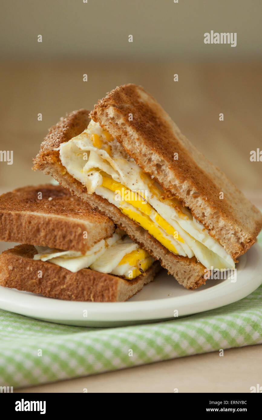 Ein Spiegelei-Sandwich Vollkorn Toast sitzt auf einem weißen Teller oben auf eine grün-weiße aufgegebenes Serviette und ein Holzbrett Stockfoto