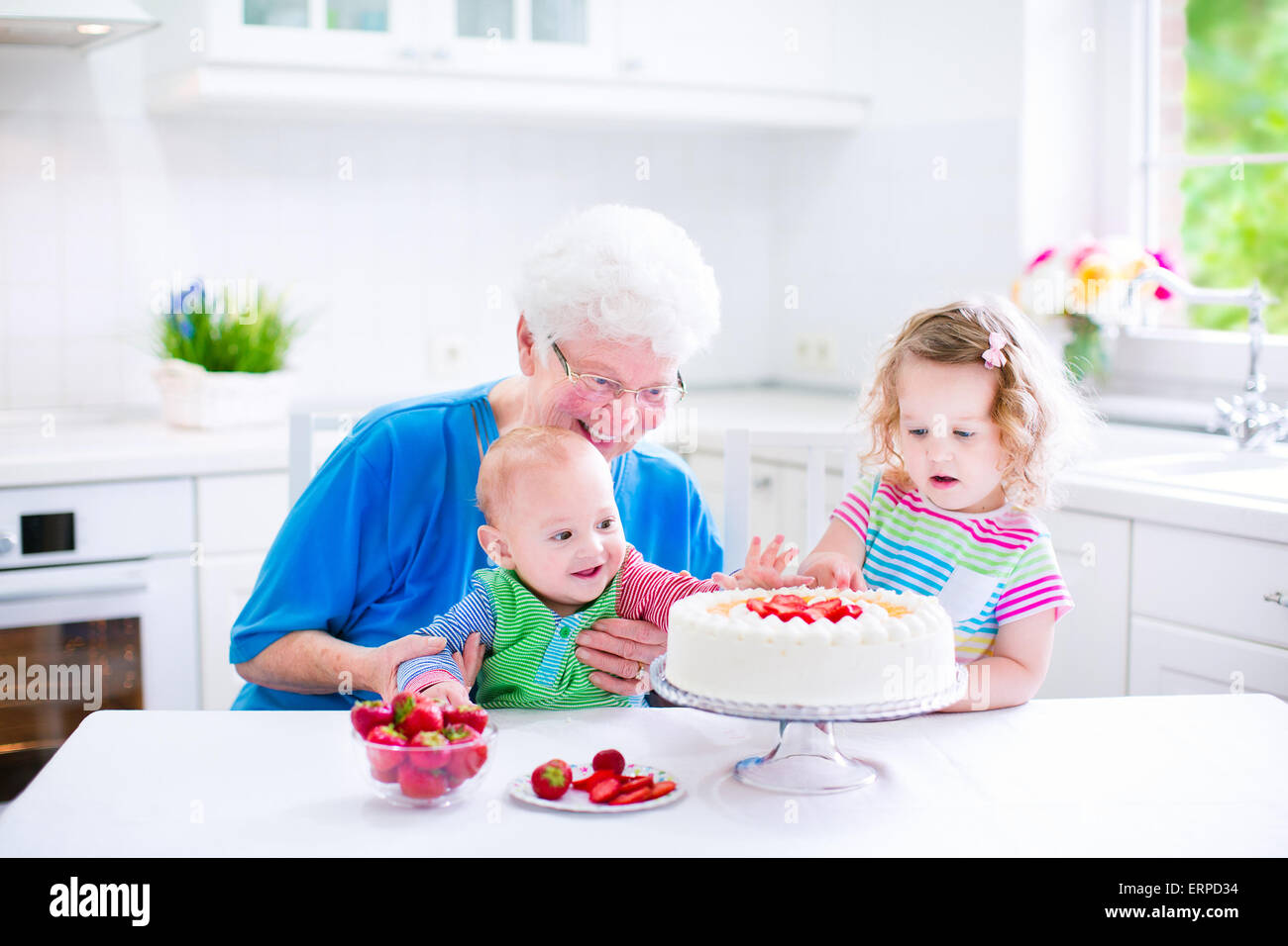 Glücklich senior Lady, liebevolle Großmutter, Backen einen hausgemachte Erdbeerkuchen mit zwei Kindern in eine weiße moderne Küche mit Fenster Stockfoto