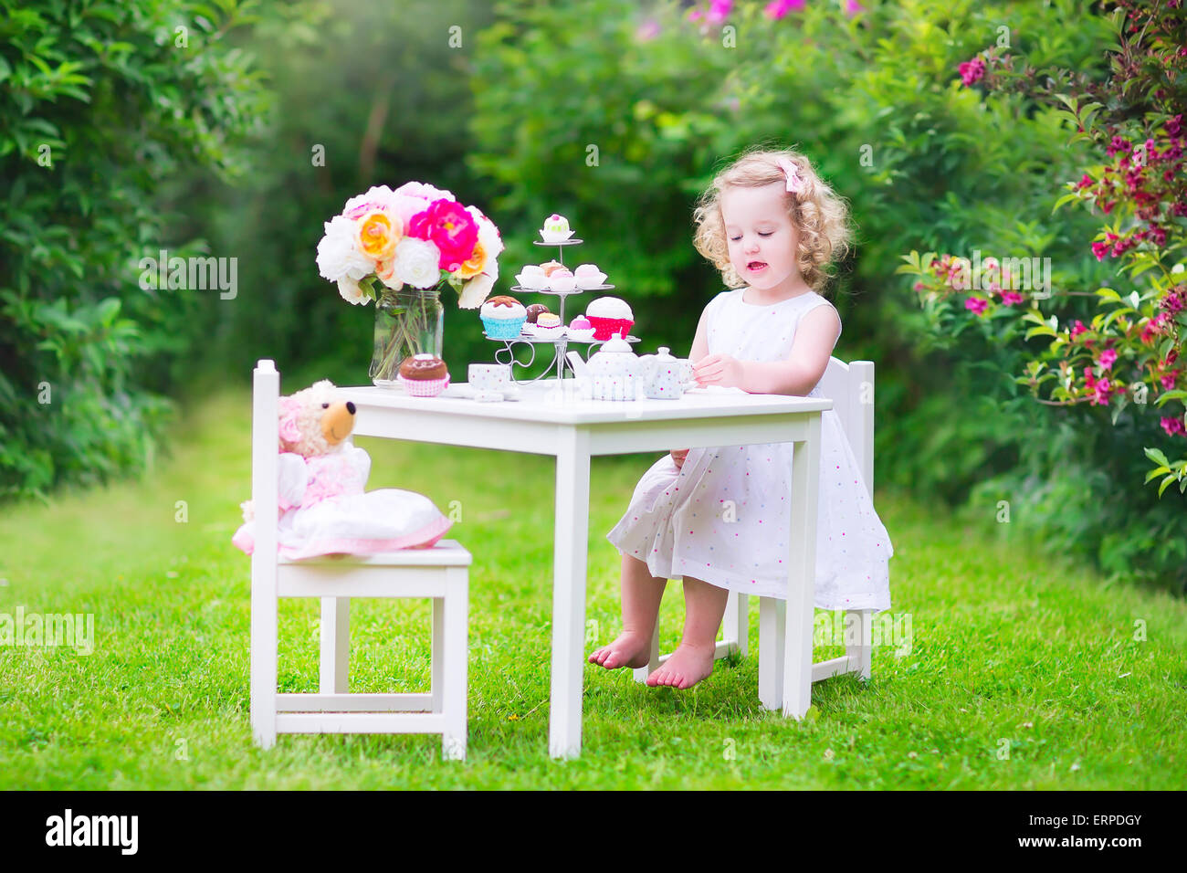Liebenswert lustige Kleinkind Mädchen mit dem lockigen Haar trägt ein buntes Kleid an ihrem Geburtstag spielen Teeparty mit einer Puppe Teddybär Stockfoto