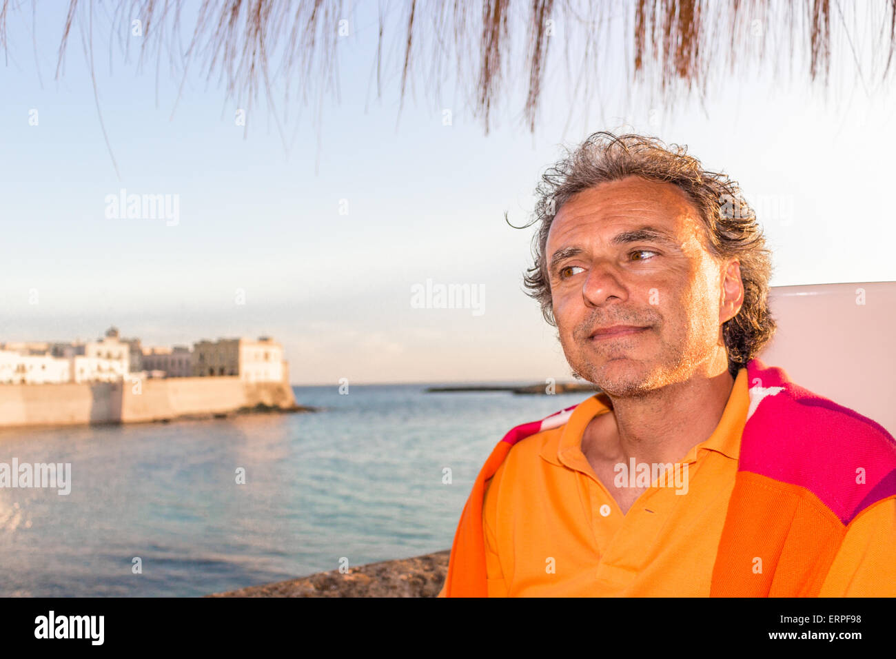 Schön gebräunte Mann mittleren Alters mit Salz Pfeffer Haar in Freizeitkleidung mit roten und orangefarbenen Pullover und gelben Poloshirt in italienischen Natur: er ist entspannt unter Dach Palm, während antike Gebäude direkt am Meer im Hintergrund stehen Stockfoto