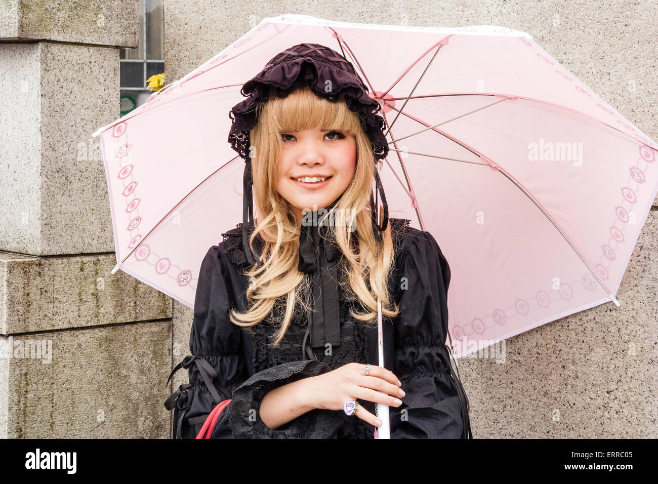 Harajuku, Tokio, eine junge japanische Frau, die sich in Lolita, rorita Fasshon, Modestil, stehend mit pinkfarbenem Regenschirm und lächelnd mit Augenkontakt bekleidet. Stockfoto