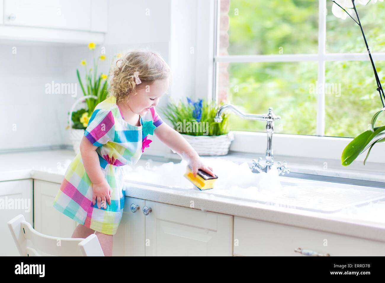 Niedliche lockige Kleinkind Mädchen in ein buntes Kleid, Abwasch, Reinigung mit einem Schwamm und mit Schaum in der Spüle in der Küche spielen Stockfoto