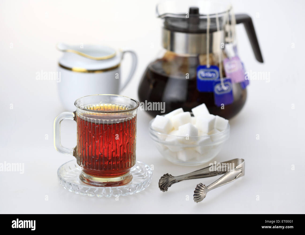 Tasse schwarzer Tee mit Würfelzucker; Milchtopf; Zangen und Wasserkocher mit Teebeuteln Stockfoto