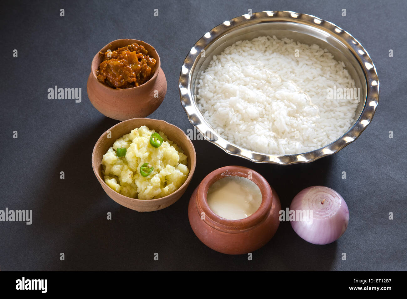 Indischer Pakhala-Reis zum Mittagessen in einer Stahlschüssel und pikante Gurkenstampfkartoffeln mit Chilis, Joghurt auf erdfarbenem Topf, Zwiebeln, schwarzem Hintergrund Stockfoto