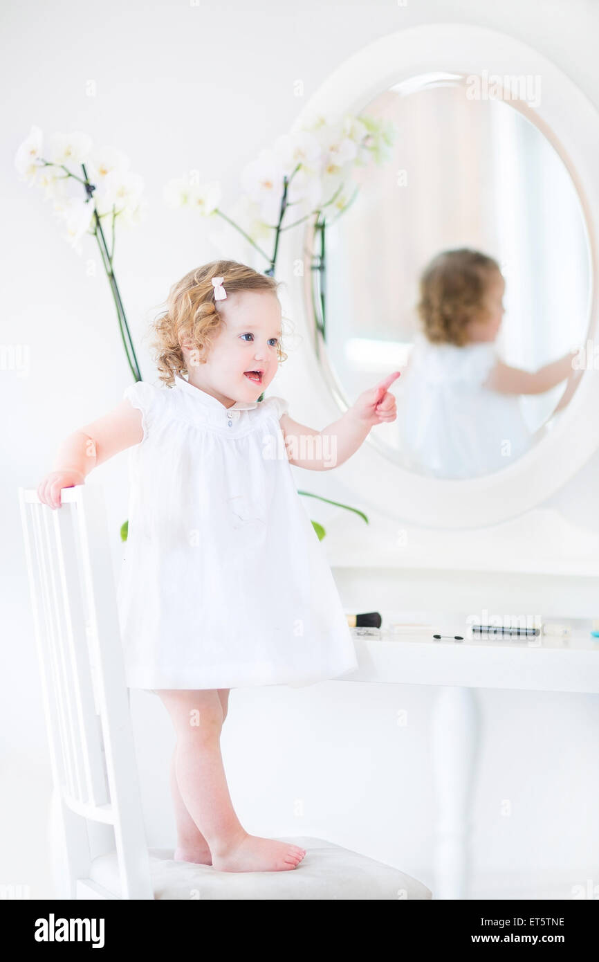 Glücklich Kleinkind Mädchen mit dem lockigen Haar trägt ein weißes Kleid mit Make up spielen und Kosmetik vor einem runden Spiegel Stockfoto