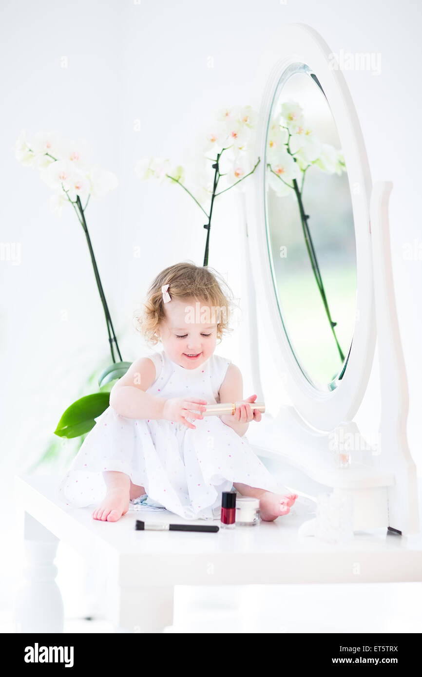 Glücklich Kleinkind Mädchen mit dem lockigen Haar trägt ein weißes Kleid mit Make up spielen und Kosmetik vor einem runden Spiegel Stockfoto